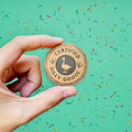 Hand holding a 'Certified Silly Goose' coin against a green background with colorful sprinkles.